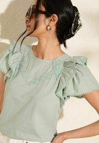 Top verde menta con maniche arricciate, caratterizzato da un bordo in pizzo decorativo attorno al collo. Tessuto morbido con una vestibilità rilassata.