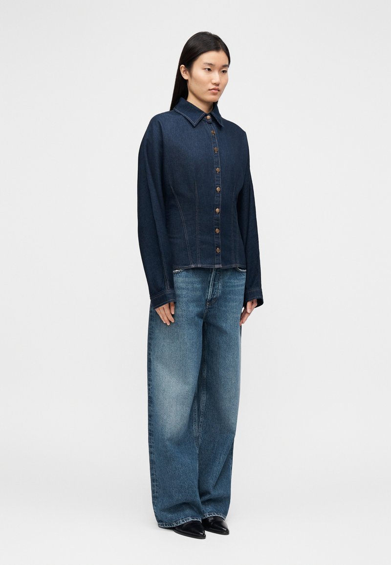 Veste en denim foncé à boutons avec un col classique, présentant un corsage ajusté, des manches longues et des boutons en bois. Associée à un jean clair ample.