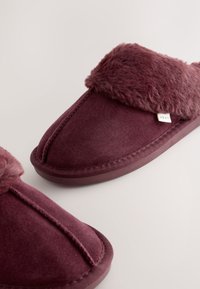Paire de chaussons d’intérieur doux de couleur bordeaux avec bordure en fausse fourrure et une étiquette en tissu sur un fond clair uni.