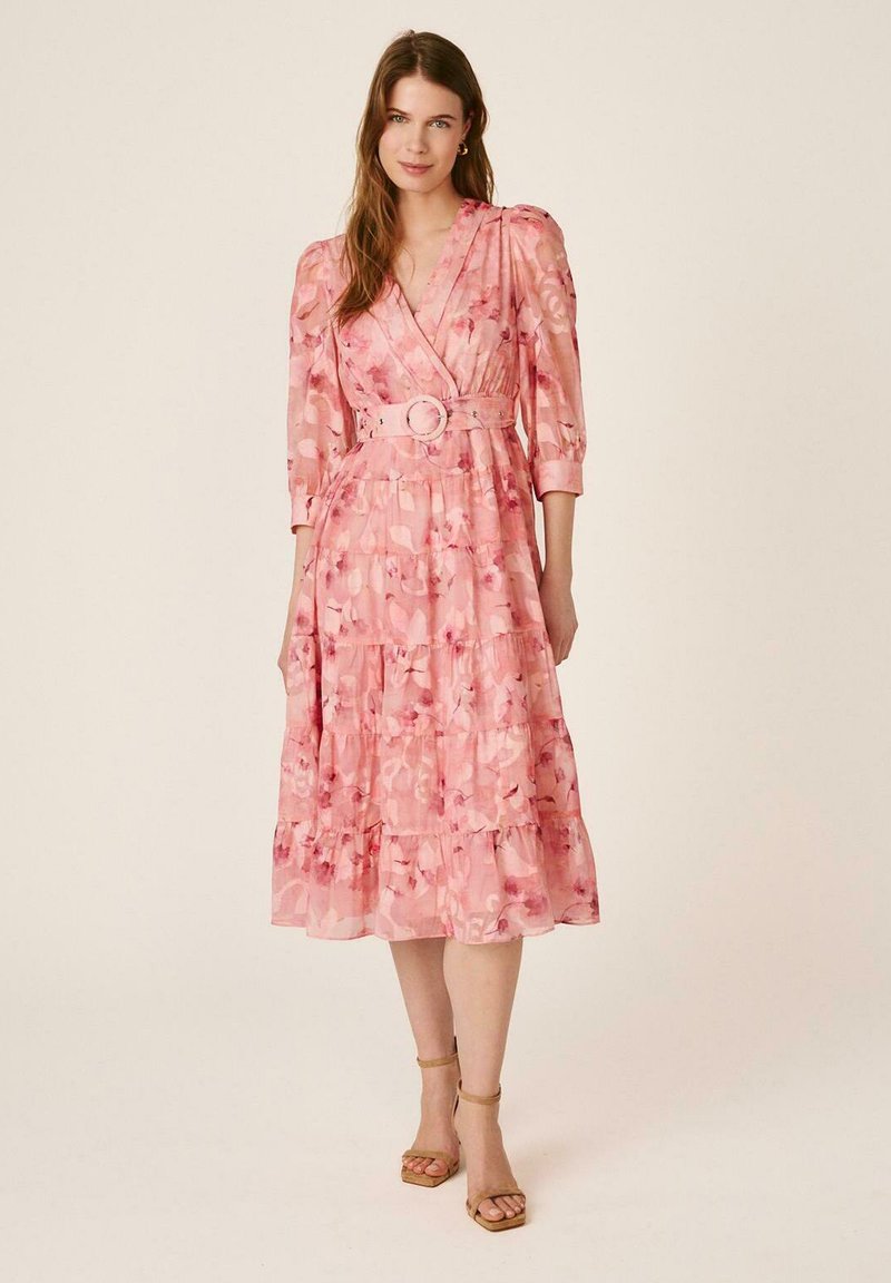 Mujer que lleva un vestido midi rosa con flores y cinturón, con mangas abullonadas y tacones marrones con tira en el tobillo, posando frente a un fondo liso.