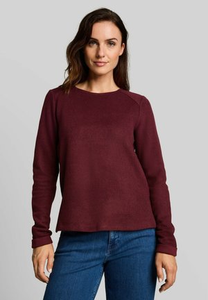 Femme aux cheveux foncés mi-longs, portant un pull bordeaux à manches longues et un jean bleu, debout devant un fond clair uni.
