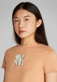 Camiseta de manga corta en color durazno, fabricada en suave algodón, que presenta un logo impreso cuadrado con texto estilizado en blanco y negro.