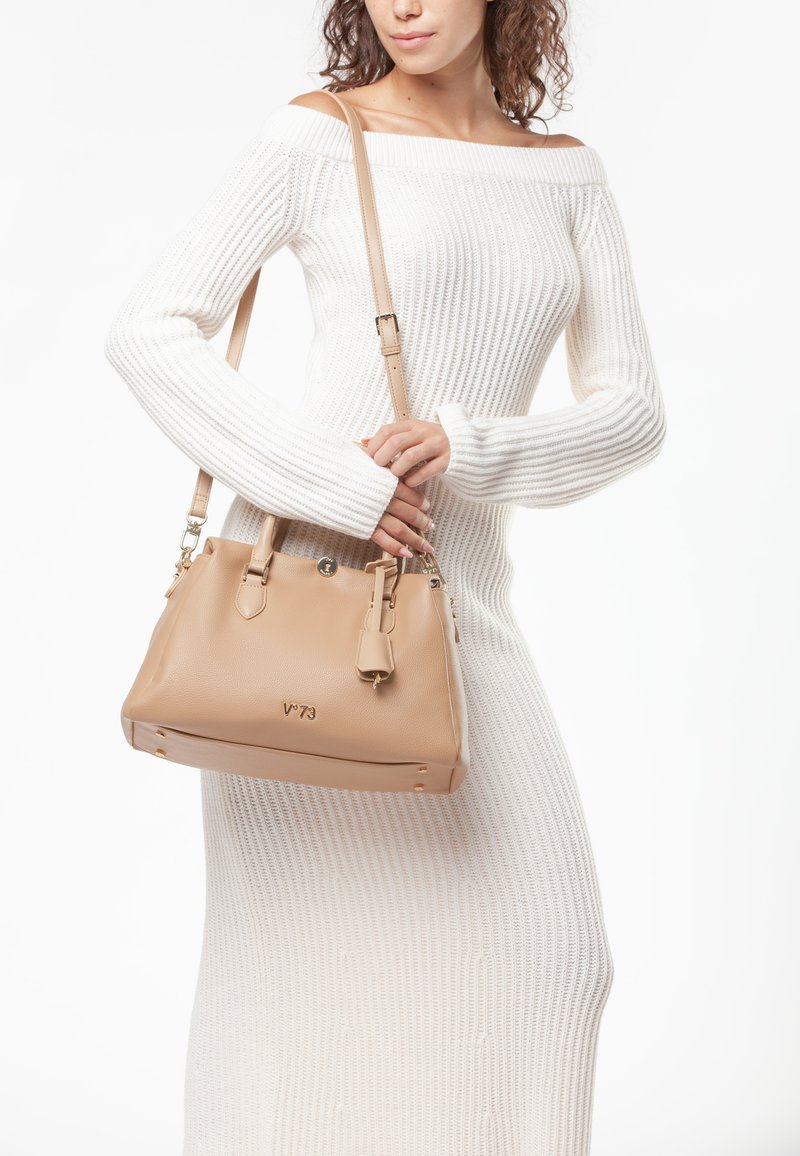 Beige leren handtas met een gestructureerd ontwerp, gouden hardware accenten, een afneembare strap en een geperst logo. Ggedragen met een gestructureerde witte trui.