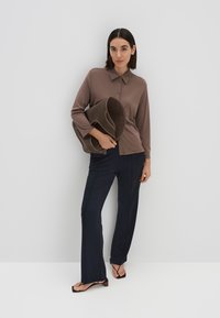 Bruine langarm polo met knopen, gecombineerd met donkerblauwe slim-fit broek. Model houdt een grote bruine geweven tas vast. Platte zwarte sandalen.