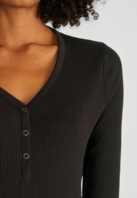 Schwarzes, geripptes Langarmoberteil mit V-Ausschnitt und drei Knöpfen am vorderen Hals. Weicher Stoff mit einer figurbetonten Passform.