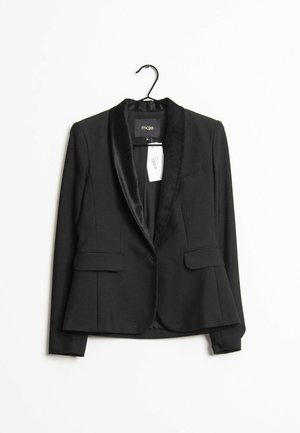 Blazer noir ajusté avec revers en satin suspendu à un cintre noir sur fond blanc, avec poches à rabat à l'avant et étiquette de prix visible.