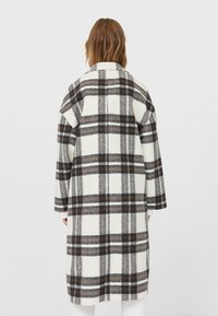 Lange geruite jas in wit, grijs en bruin, gemaakt van zacht, textuurstof. Heeft een notch-kragen en een verlaagde schouderlijn.