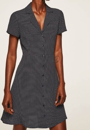 Kobieta ubrana w czarną, krótkorękawową sukienkę na guziki w białe kropki z dekoltem w serek, stojąca z jedną ręką przy udzie.