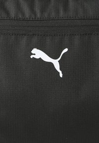 Tissu noir texturé avec des rayures horizontales, arborant un logo Puma blanc. Le design comprend une fermeture éclair sécurisée en haut.