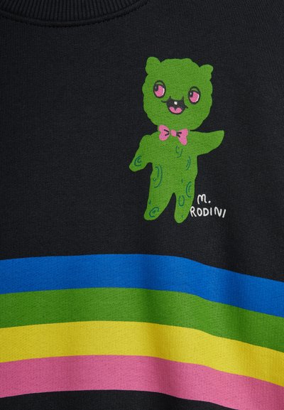 Πράσινος καρτουνίστικος αρκούδος με ροζ μάτια και παπιγιόν πάνω από οριζόντιες ρίγες σε μπλε, πράσινο, κίτρινο κ αι ροζ σε σκούρο ύφασμα.