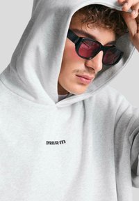 Haut de survêtement gris clair avec une capuche à cordon de serrage, doté d'un petit logo noir sur la poitrine. Porté avec des lunettes de soleil à monture noire avec des verres rougeâtres.