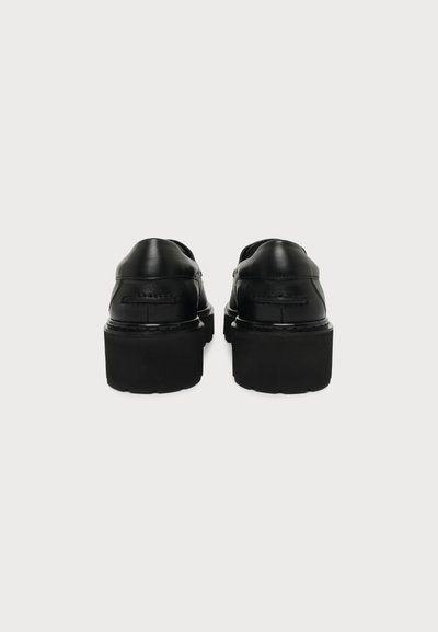 Paire de chaussures à plateforme en cuir noir avec semelles épaisses vues de dos sur un fond uni clair.