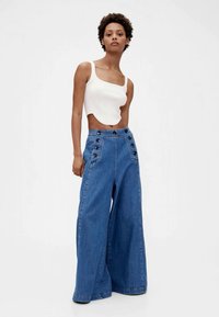 Wide-leg denim trousers with high waist and button accents, paired with a fitted white crop top. The fabric is blue and textured.