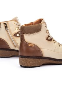 Bottines à lacets en cuir beige avec des accents marron. Présentent une semelle extérieure texturée et des coutures décoratives. Fermeture éclair sur le côté pour un enfilage facile.