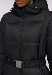 Svart puffjacka med dragkedja fram, elastisk midja och justerbar huva. Har rektangulär sömnad för textur och en metallspänne.