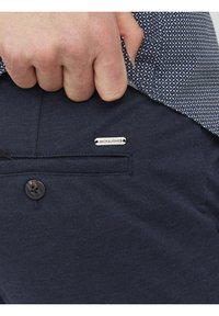 Námornícko modré kalhoty s knoflíkovým zapínáním, detaily kapes a stříbrnou kovovou štítkem se logem "JACK & JONES" vyšitém na boku.