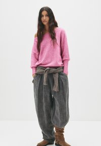 Roze oversized gebreide trui met geribde mouwen, gecombineerd met grijze wijde broekspijpen die met een bruine gestructureerde riem op de taille zijn vastgebonden. Bruine kniehoge laarzen.