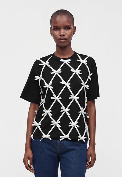 Personne portant une chemise noire à manches courtes décorée d’un motif croisé blanc de nœuds noués, associée à un jean bleu sur un fond uni.