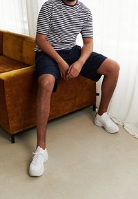 Muž v pruhované košili s krátkým rukávem, námořnických šortkách a bílých teniskách sedí na hořčicově zbarvené pohovce v jasné místnosti.