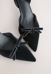 Paire de ballerines en velours noir à bout pointu avec un détail fin de nœud sur le devant, sur un fond clair.