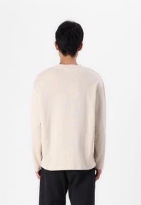 Chemise beige clair à manches longues avec une coupe décontractée, présentant un col rond, des épaules tombantes et une texture de tissu douce et lisse.