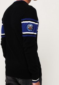Uomo che indossa un maglione nero con strisce blu e bianche sulla parte superiore del braccio e una toppa rotonda a tema montano sulla manica.