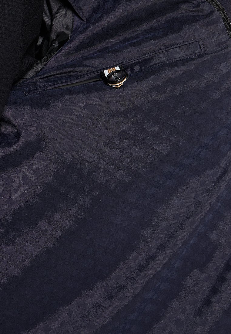 Intérieur d'une veste marine avec des motifs géométriques subtils, texture lisse, et une poche avec une fermeture à bouton et une sangle décorative.