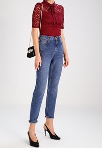 Burgunder Spitzenoberteil mit kurzen Ärmeln und Schleife, kombiniert mit hoch geschnittenen blauen Jeans. Schwarze spitze Absätze und kleine schwarze Handtasche.