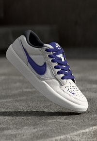 Nike tenisky s šedým koženým svrškem, fialovými akcenty, černým jazykem, perforovanou špicí a texturovanou bílou gumovou podrážkou.