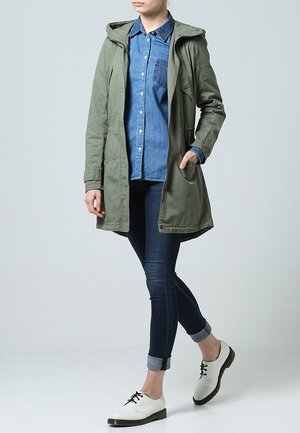 Zelená bunda s kapucí, džínová košile, tmavé úzké džíny a bílé boty s černou podrážkou. Má kapsy a neformální střih.