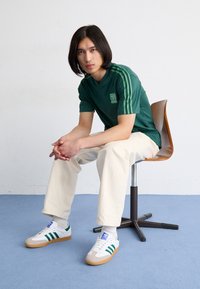 Grön v-ringad t-shirt med tre gröna ränder på ärmarna, i kombination med krämfärgade byxor och vita sneakers med gr öna och blå detaljer.