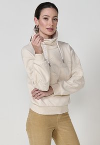 Hoodie de couleur crème avec un col haut, présentant une broderie florale texturée, des cordons de serrage à bascule et des poignets côtelés. Porté avec un pantalon beige.