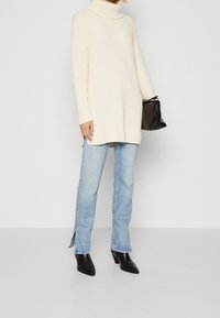 Cremfarbener gerippter Rollkragenpullover, kombiniert mit hellblauen Jeans mit seitlichen Schlitzen und schwarzen Lederstiefeln mit Absatz. Schwarze Tasche in der Hand.