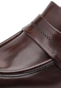 Gros plan d’une chaussure habillée en cuir marron montrant une finition lisse, des détails de couture et une lanière en cuir sur le dessus.