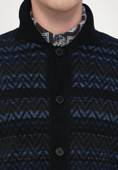 Vilnas kardigāns ar zigzaga rakstu melnā un zilā krāsā, ribotu apkakli un piecām melnām pogām priekšpusē.
