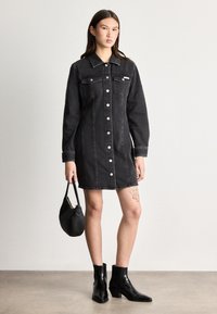 Vestido de ganga preto com dois bolsos no peito, botões prateados e colarinho. Mangas longas e silhueta ajustada. Botas de tornozelo pretas e bolsa.