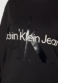 Černá mikina s lesklým černým potiskem, na kterém je velké "CK" a text "Calvin Klein Jeans" v bílé, kulatý výstřih, měkký materiál.