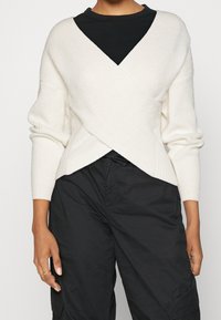 Vit stickad omslagströja med ribbat mönster, långa ärmar, djup V-ringning, lagd över en svart topp, ihop med svarta cargobyxor.