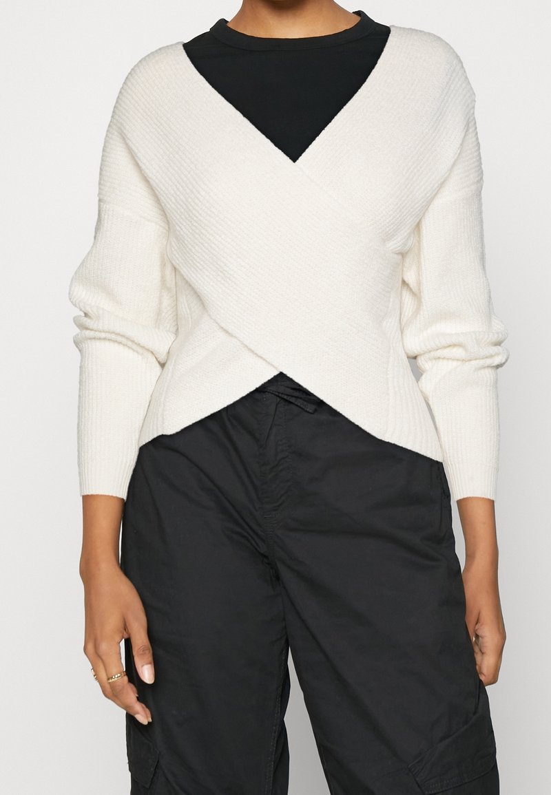 Vit stickad omslagströja med ribbat mönster, långa ärmar, djup V-ringning, lagd över en svart topp, ihop med svarta cargobyxor.