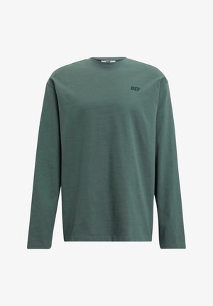 Chemise à manches longues en tissu doux de couleur vert muted. Présente un col rond et un petit logo ton sur ton "DEF" sur la poitrine. Design simple.