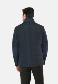 Giacca trapuntata navy con colletto alto, toppe sui gomiti strutturate e dettagli di cucitura visibili. Tessuto liscio con una vestibilità strutturata.