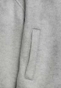 Pelēkas sajūtu audums ar sānu šuvi, kas izceļas ar taisnstūra formas kabatu un redzamām šuvēm, nodrošinot mīkstu tekstūru un minimālisma dizainu.