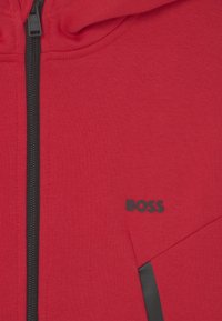 Sweat à capuche rouge avec fermeture éclair noire, comportant un logo noir. Texture douce avec une fermeture éclair à l'avant et un détail de poche inclinée.