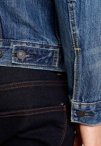 Džínová bunda s modrým efektem praní, zdobená kovovými knoflíky a žlutým prošitím, v kombinaci s tmavými džínami s oranžovým prošitím.