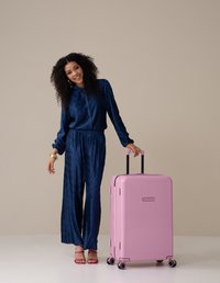 Roze harde koffer met verticale groeven, zwarte uitschuifbare handgreep en wielen. Model in een marineblauw gestructureerd jumpsuit staat ernaast.