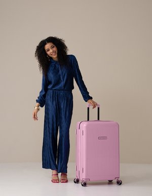 Roze harde koffer met verticale groeven, zwarte uitschuifbare handgreep en wielen. Model in een marineblauw gestructureerd jumpsuit staat ernaast.