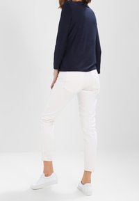 Kvinna klädd i en marinblå långärmad topp, vita slim-fit byxor och vita sneakers, står med ett ben något böjt mot en enfärgad bakgrund.