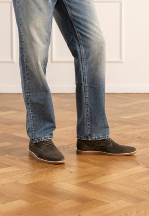 Gambe con jeans blu sbiaditi e scarpe stringate in camoscio grigio scuro, in piedi su un pavimento in legno con motivo, all'interno di un ambiente.
