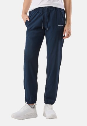 Person iført navyblå joggers med snøre i taljen, elastiske manchetter og hvide atletiske sneakers, stående på en hvid baggrund.