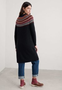 Černé pletené šaty s barevným vzorem na výstřihu, dlouhými rukávy a volným střihem. Kombinované s džíny se zahnutými okraji a dřeváky.